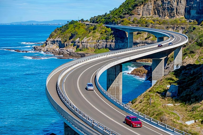 Curved elevated highway bridge over turquoise ocean water with rocky cliffs and green vegetation on a coastal landscape