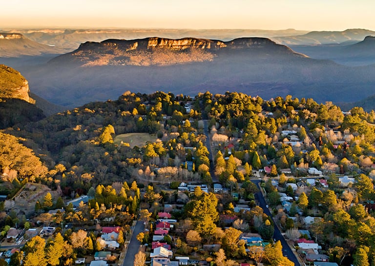 Aerial view of a residential suburb nestled in forested hills with mountain cliffs visible in misty distance at sunrise